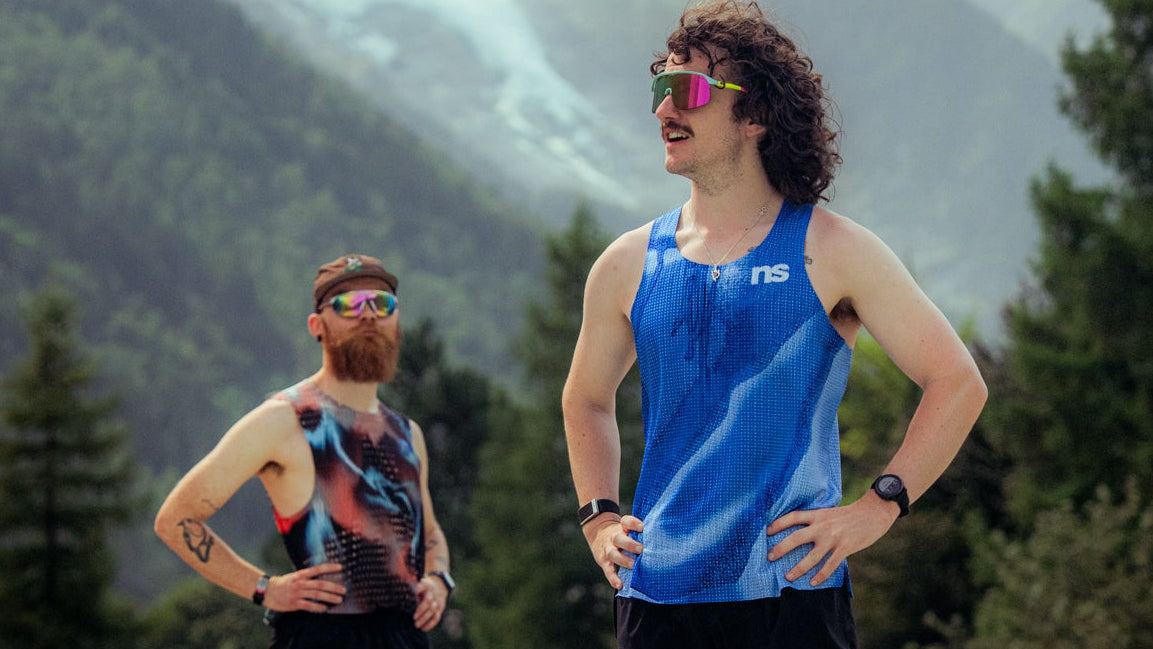 Two men in athletic wear standing on an outdoor track field with mountains and trees in the background. The man in the foreground wears a blue sleeveless athletic top, black shorts, white socks, and white running shoes, while the man behind him wears a multicolor top, black shorts, and colorful running shoes.